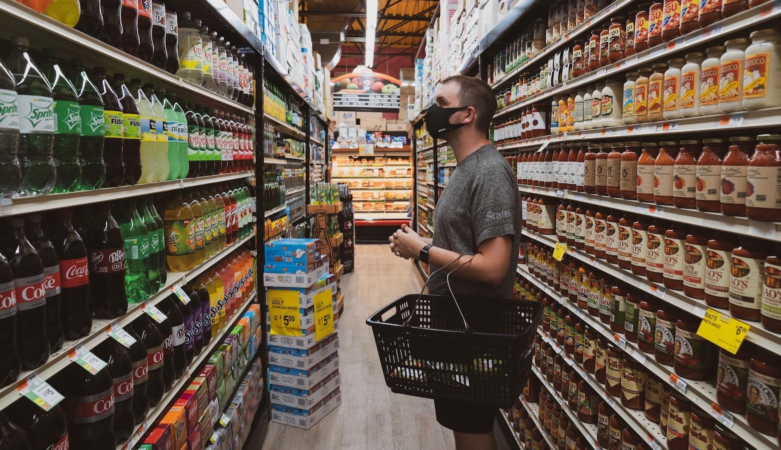 一名戴着布口罩、穿着灰色衬衫的年轻人站在一家杂货店里，前臂上放着一个购物篮。他正在看满架子的软饮料。